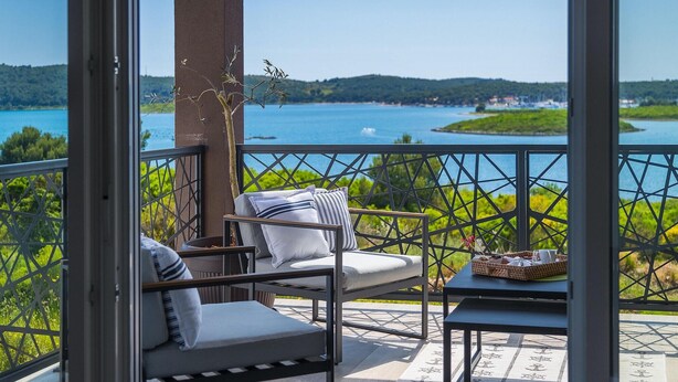 Ein Balkon mit einer Couch, einem Sessel, einem kleinen Tisch und Blick auf einen See und Grünflächen.