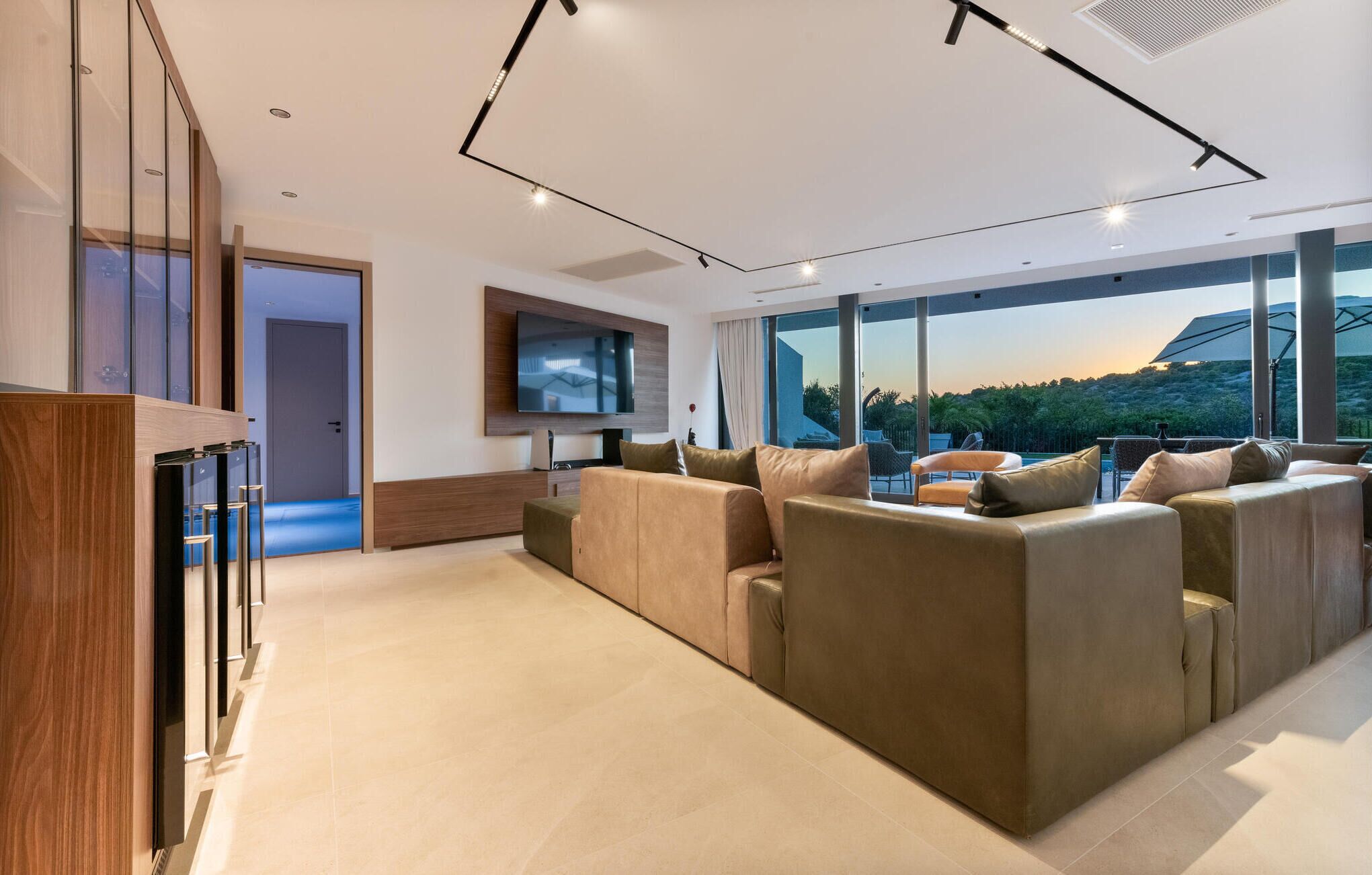 A modern living room with a large sectional sofa, a flat-screen TV mounted on the wall, and a large window offering a view of the outdoors.