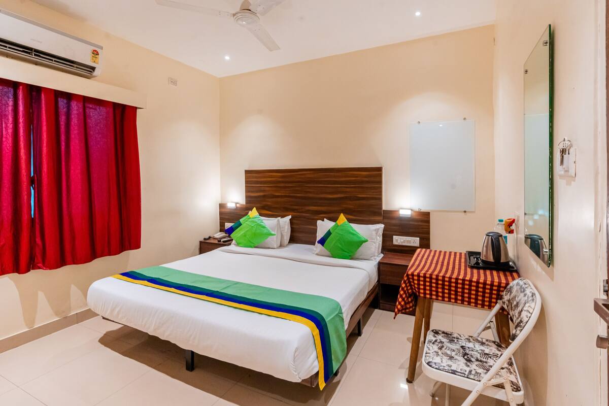 A hotel room with a bed, a desk with a chair, a mirror, and a red curtain.