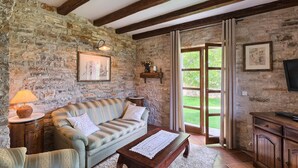 Una habitación rústica con paredes de piedra vista, un banco de madera, un sofá con cojines, una mesita y un televisor sobre un mueble de madera.