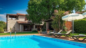 Una piscina con sillones de descanso y sombrillas, un edificio de piedra con techo de tejas y abundante vegetación.