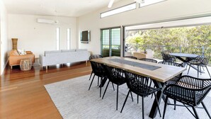 Una sala de estar moderna con una mesa de comedor de madera, sillas negras, un sofá gris y un amplio ventanal con vistas a zonas verdes.