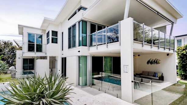 Una casa moderna de dos pisos con un amplio balcón de vidrio, piscina y un jardín bien cuidado.