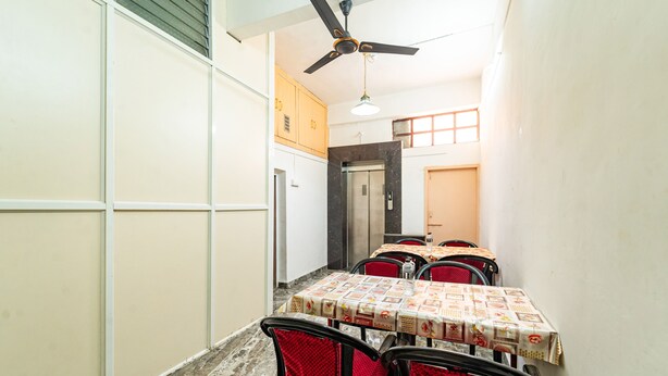 A dining area with a table set for four, red chairs, a ceiling fan, and a view of a corridor with a door and an elevator. - Itsy Hotels Mansi (Nashik)