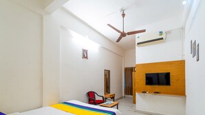 A hotel room with a bed, a TV mounted on the wall, an air conditioning unit, a ceiling fan, and a small table with a chair. - Itsy Hotels Mansi (Nashik)