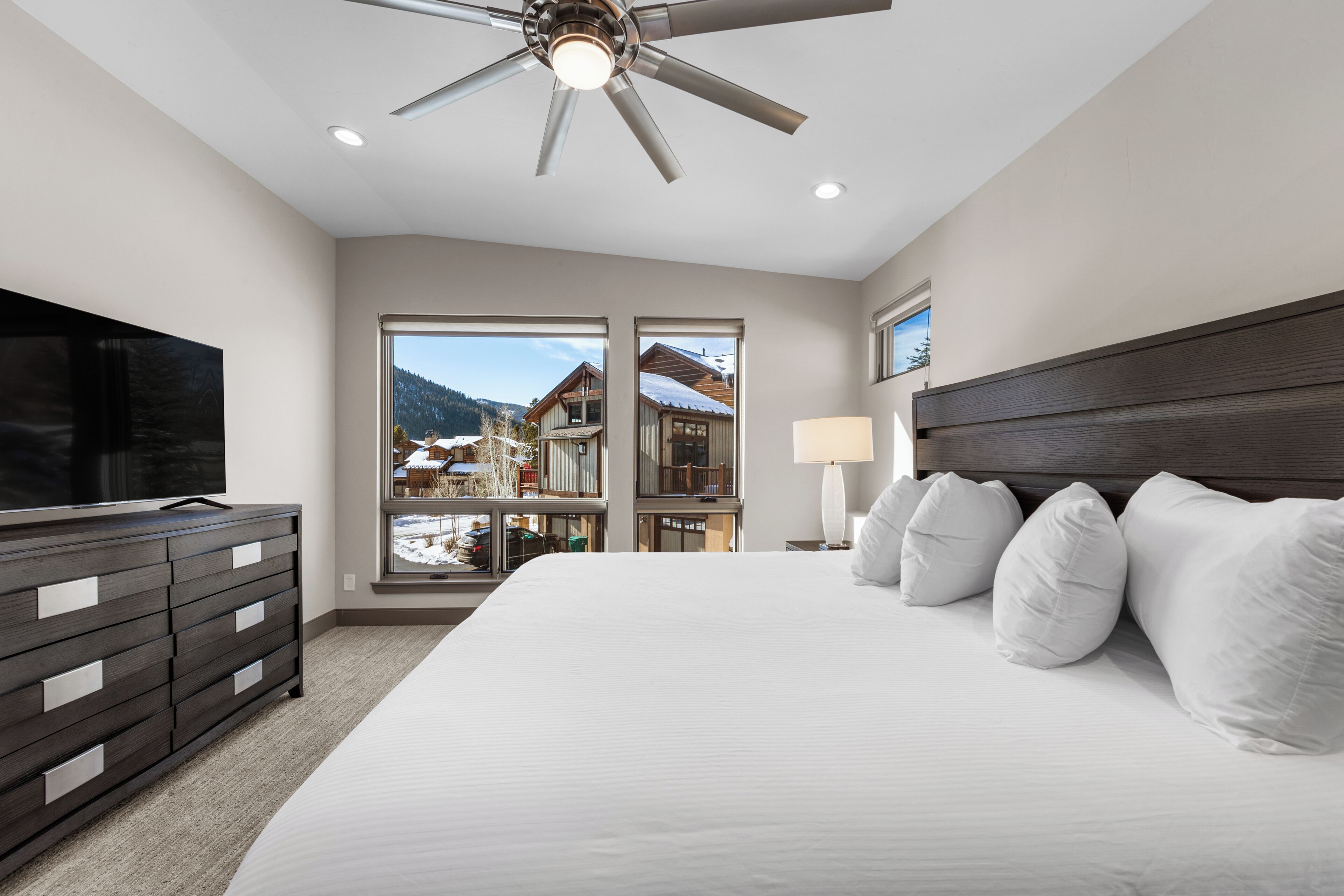 A bedroom with a large bed, a TV, a ceiling fan, and a view of a snowy mountain.