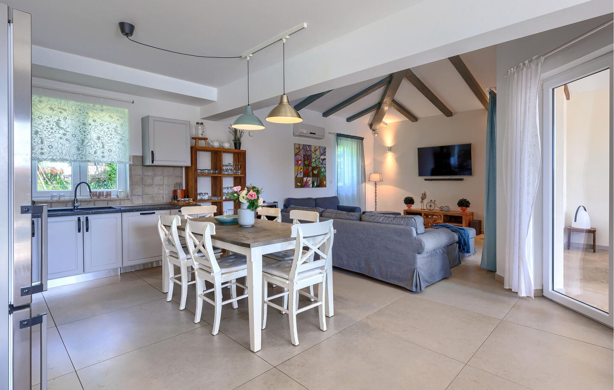 A modern kitchen with white cabinets, a dining table with white chairs, a grey sofa, and a TV mounted on the wall.