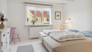 Et soveværelse med seng, kommode, stol, et vindue med udsigt til bygninger og en vægmonteret lampe.