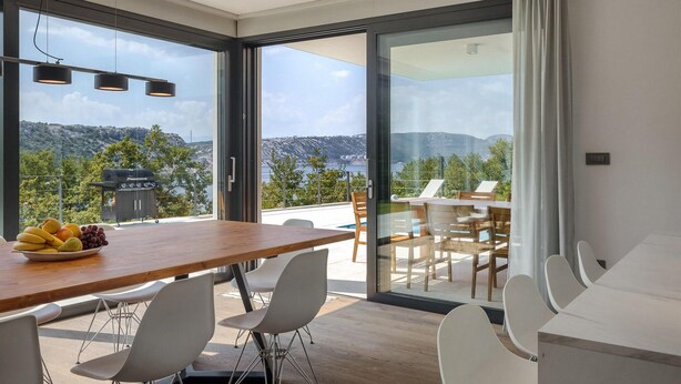 Et moderne spiseområde med et træbord og hvide stole, en grill og udsigt til et bjergrigt landskab gennem store glasdøre.