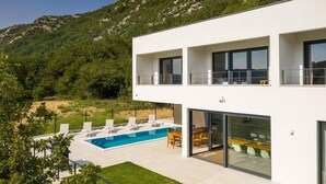 Et moderne hus med to etager, en swimmingpool, udendørs møbler og udsigt til et bjergrigt landskab.