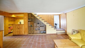 Un amplio espacio habitable con una escalera de madera que lleva a un altillo, una zona de cocina con botellas de vino y un sofá amarillo.