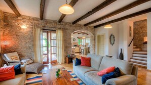 Una sala de estar acogedora con pared de piedra, vigas de madera, un sofá con cojines coloridos, una silla de mimbre y una mesa de centro de madera.