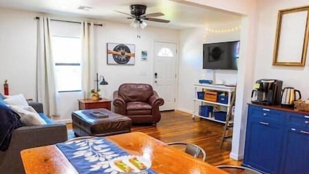 Una sala de estar con un sofá de cuero marrón, una mesa de comedor de madera con camino de mesa azul, un ventilador de techo, un televisor en la pared y una zona de cocina con armarios azules.