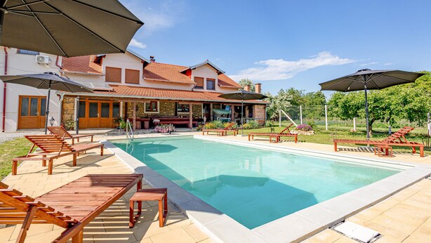 Un área de piscina con sillones reclinables y sombrillas, una casa de piedra con techo de tejas y un cielo azul despejado.