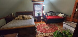 A bedroom with two beds, a wooden headboard, a red and white patterned rug, a small table with a lamp, and a window with blinds.