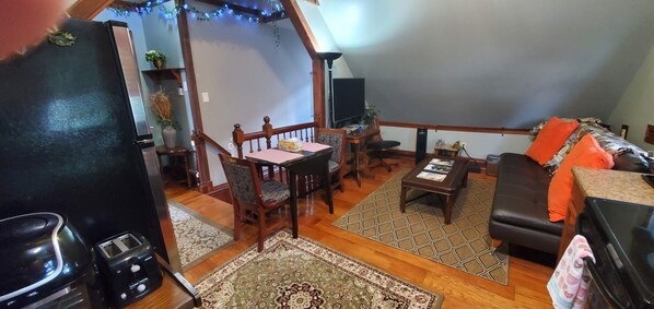A cozy living room with a wooden dining table, chairs, a sofa, and a coffee table. There is a TV, a speaker, and a bookshelf with plants.
