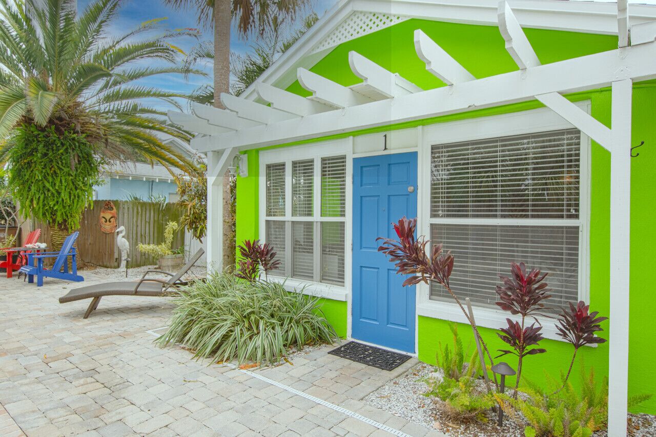A bright green and white house with a blue door, a porch with a white railing, and a small garden with plants and a palm tree.