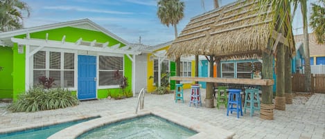 Uma área de piscina com uma estrutura de telhado de palha, um edifício verde com uma porta azul e banquinhos coloridos.