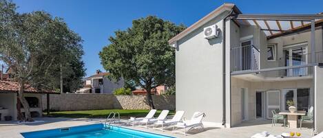 Una piscina con sillas de descanso, un muro de piedra y un edificio de dos plantas con balcones.
