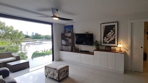 Ein modernes Wohnzimmer mit großem Fernseher, Deckenventilator, Balkon mit Blick auf einen Fluss und Gebäude sowie einem Bücherregal.