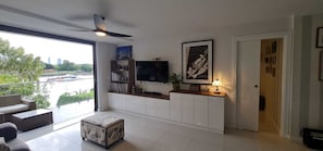 A modern living room with a large TV, a ceiling fan, a balcony with a view of a river and buildings, and a bookshelf.