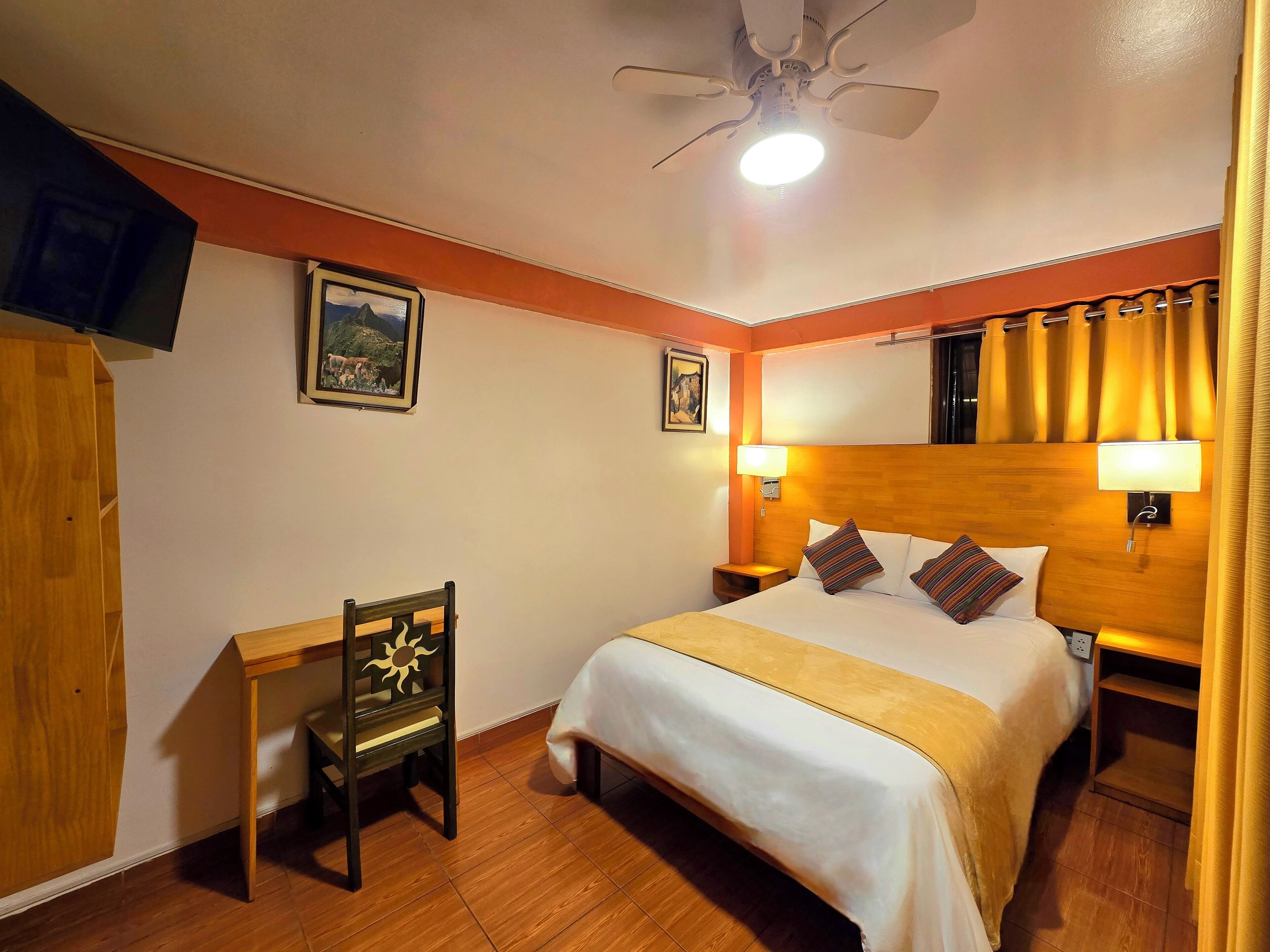 Une chambre d’hôtel équipée d’un lit, d’un bureau, d’une chaise, d’un ventilateur de plafond et de deux lampes fixées au mur.