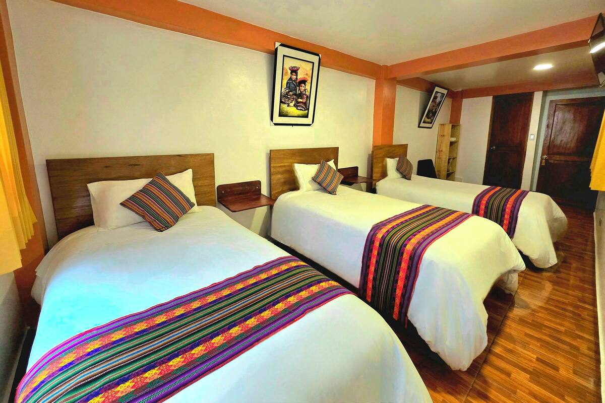 A hotel room with three single beds, each with a wooden headboard and white bedding. There is a framed picture on the wall above the beds, and a wooden door to the right.