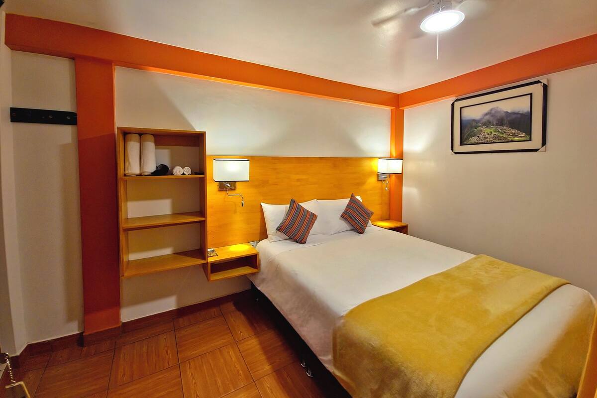 A hotel room with a wooden headboard, a bedside table with a lamp, a shelf with towels, and a bed with a yellow blanket.
