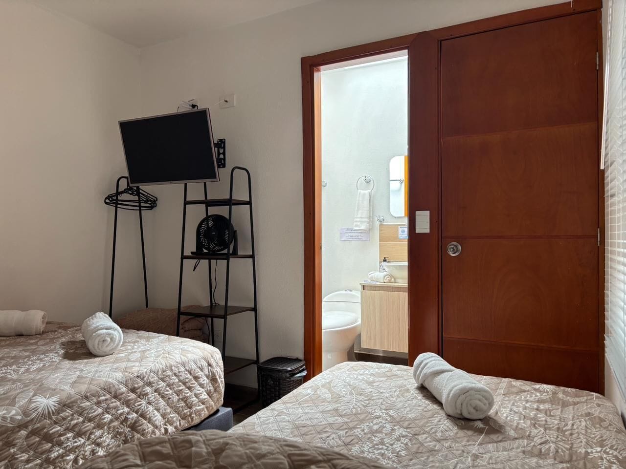 Habitació d'hotel amb dos llits, un televisor sobre un suport, un armari de fusta i un bany visible a través d'una porta oberta.