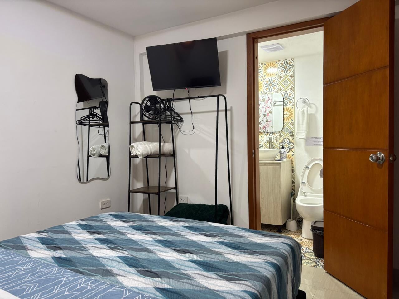 Un dormitorio con cama, televisor montado en la pared, un tendedero y un baño visible a través de una puerta abierta.