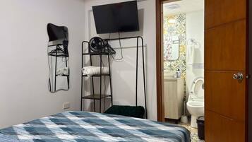 Un dormitorio con cama, televisor montado en la pared, un tendedero y un baño visible a través de una puerta abierta.