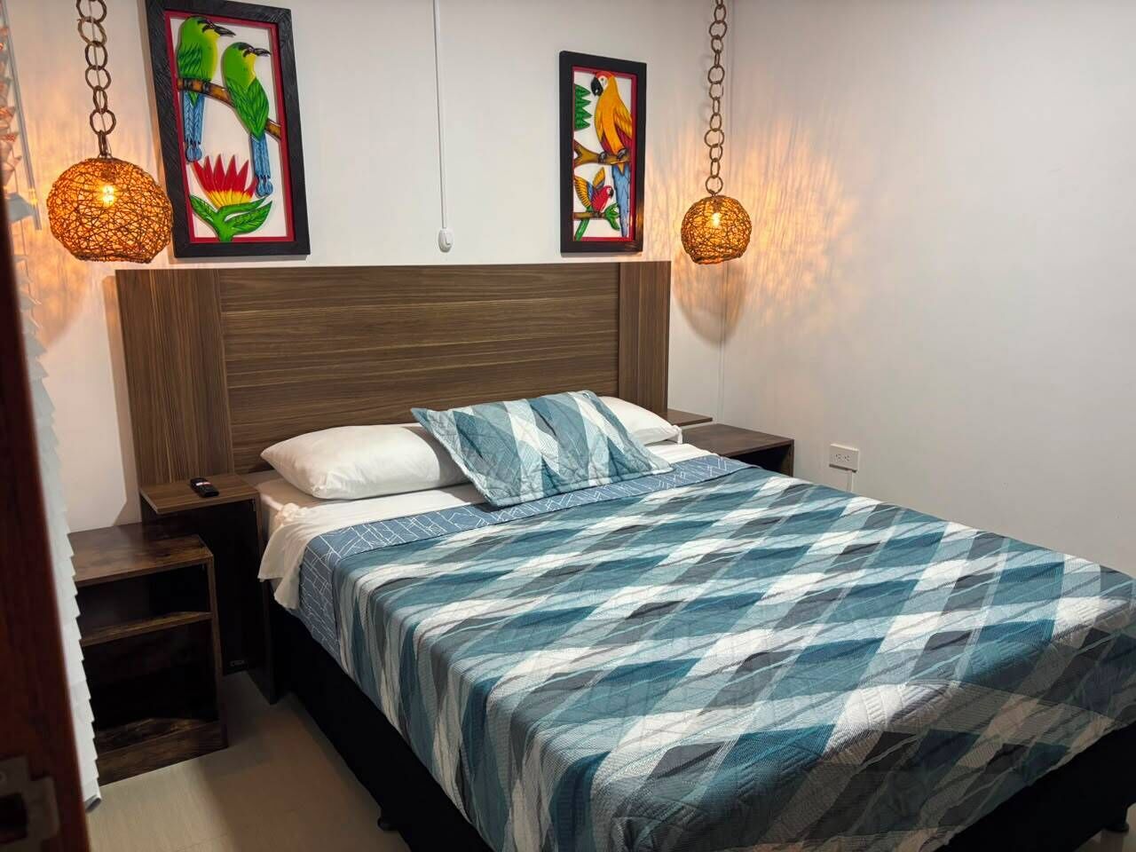 Uma cama com colcha azul e branca xadrez, cabeceira de madeira, duas luminárias fixadas na parede e duas pinturas emolduradas de pássaros.