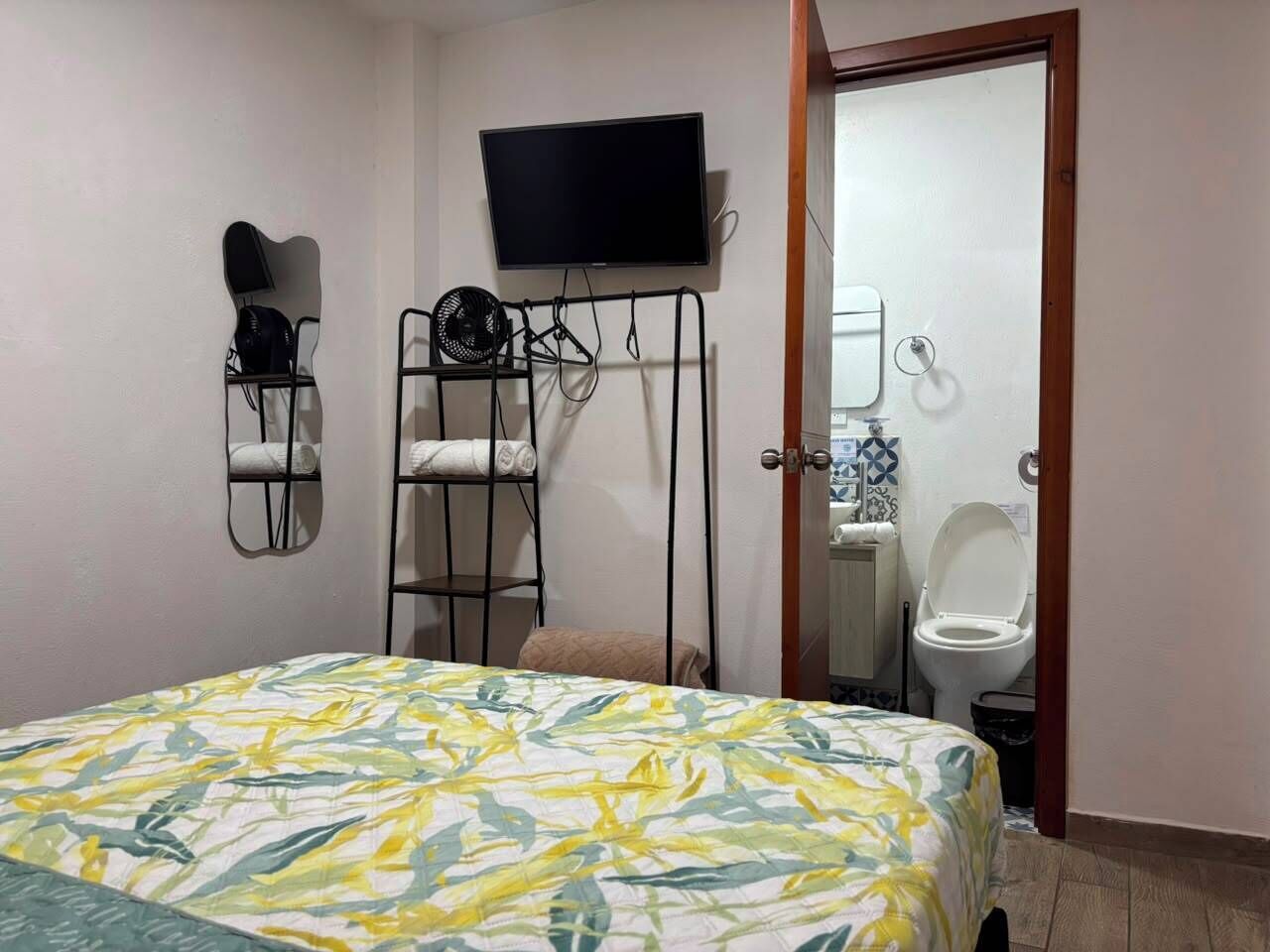 Een slaapkamer met een bed, een aan de muur bevestigde televisie, een ventilator en een zichtbare badkamer door een geopende deur.