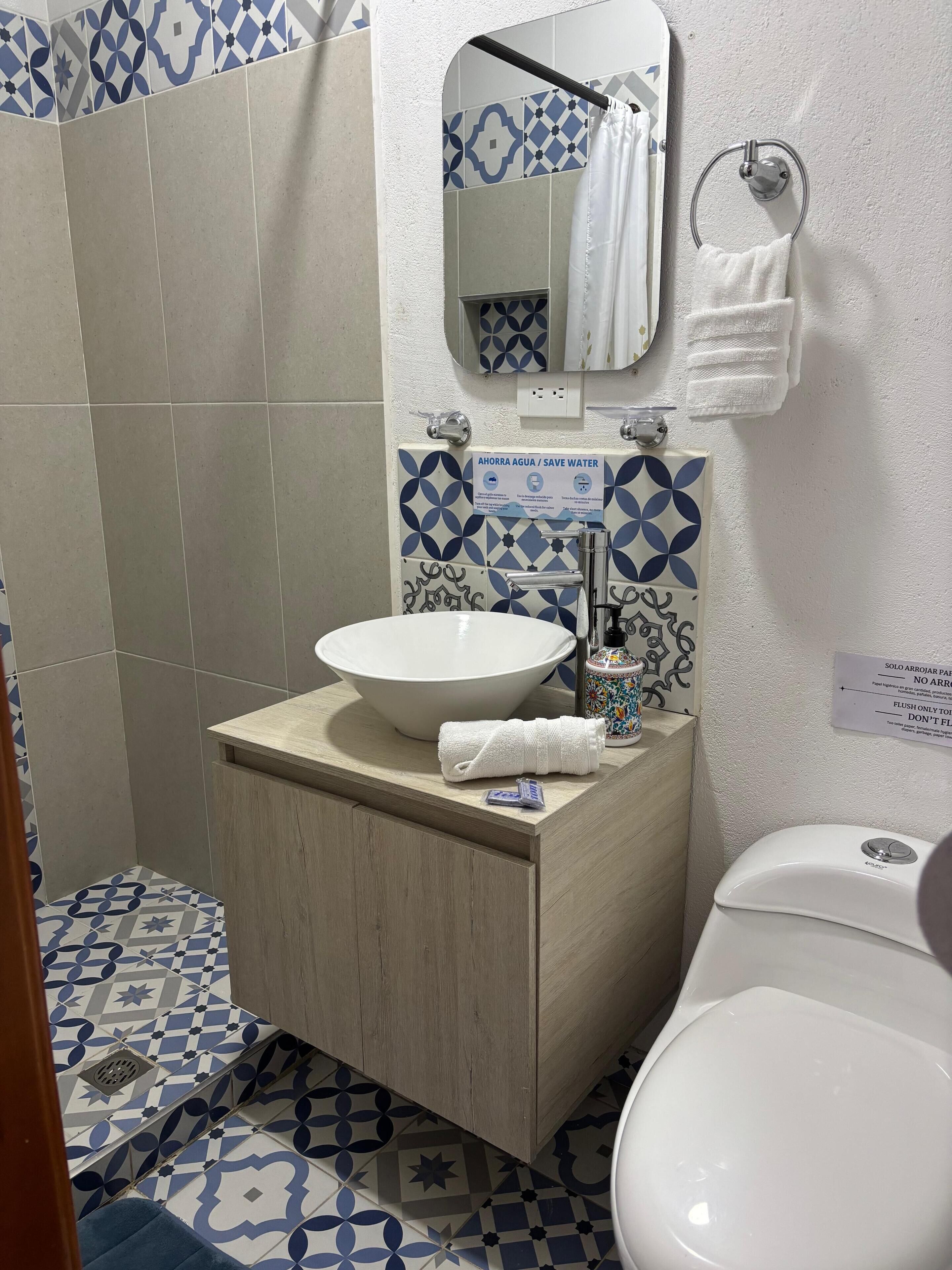 Casa de banho com lavatório branco, espelho e sanita. Há um aviso na parede que diz 'AHORRA AGUA / SAVE WATER'.