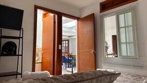 Un dormitorio con cama, una puerta de madera, una ventana con persianas y vista a un comedor.