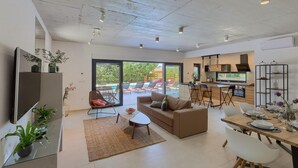 Un salon moderne comprenant un canapé, une table à manger pour quatre personnes, un coin cuisine avec des tabourets de bar et une grande porte-fenêtre coulissante donnant sur un espace extérieur aménagé pour se détendre.