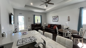 Una cocina moderna con una isla central, una zona de comedor con sillas blancas, un sofá de cuero oscuro y un ventilador de techo.