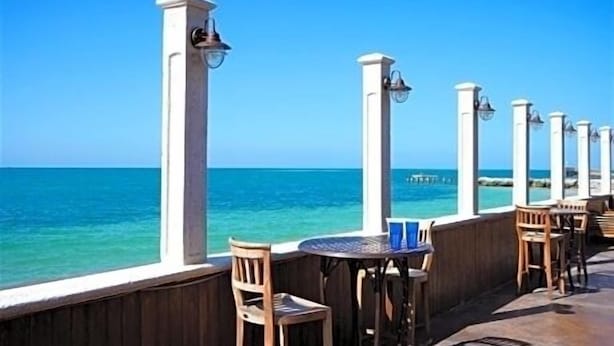 Una terraza frente al mar con sillas y mesas de madera, pilares blancos con lámparas y vista al océano.