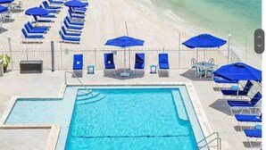 Una piscina rectangular con solárium empedrado, rodeada de sillas reclinables y sombrillas azules.