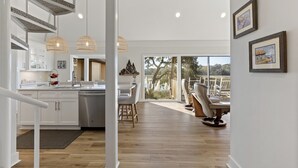 A modern kitchen with white cabinets, a dining area with a large window, and a staircase with a unique design. - Beautiful Salt Marsh Cottage in Moss Creek, where serenity awaits. (Hilton Head Island)