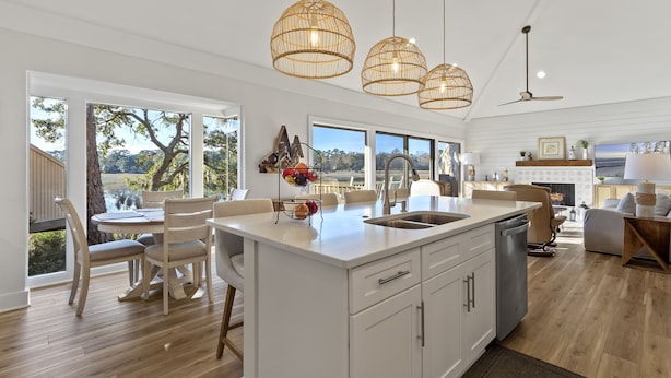 A spacious kitchen with a large island, white cabinetry, and a view of the outdoors. There are three hanging pendant lights and a ceiling fan. - Beautiful Salt Marsh Cottage in Moss Creek, where serenity awaits. (Hilton Head Island)