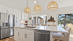 A modern kitchen with white cabinetry, stainless steel appliances, and a large island with a sink. There are three wicker pendant lights above the island. - Beautiful Salt Marsh Cottage in Moss Creek, where serenity awaits. (Hilton Head Island)