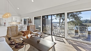 A spacious living area with a large window overlooking a deck and water. There is a leather chair, a wooden table, and a hanging light fixture. - Beautiful Salt Marsh Cottage in Moss Creek, where serenity awaits. (Hilton Head Island)