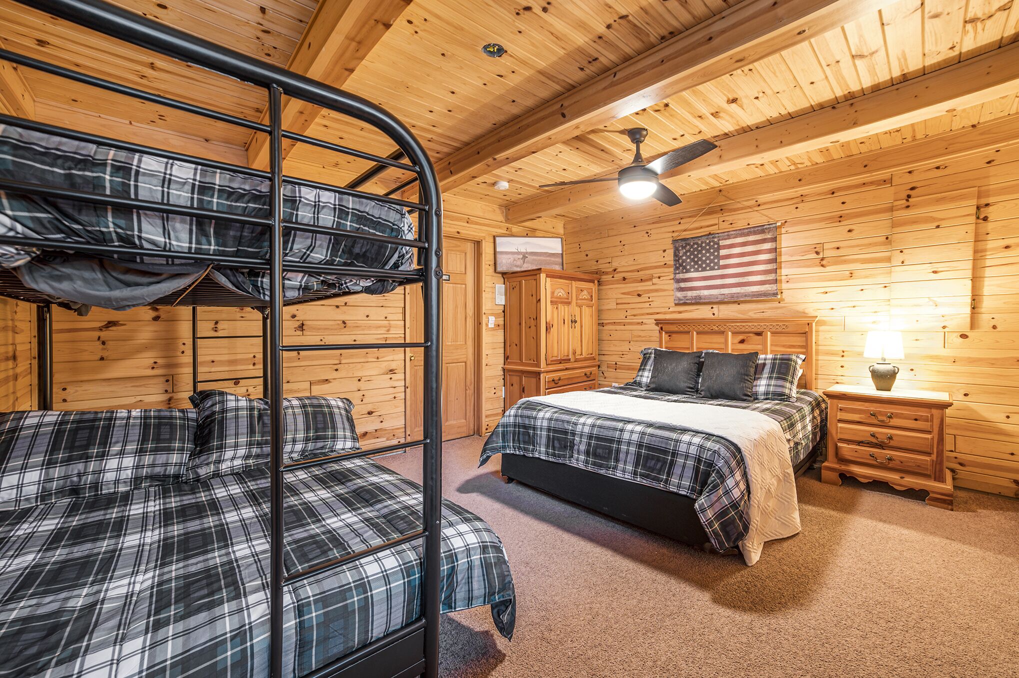 Quarto estilo cabana com beliches, uma cama de solteiro, paredes de madeira, uma bandeira americana, um ventilador de teto e uma mesa de cabeceira de madeira com uma luminária.