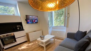 A modern living room with a grey sofa, a white coffee table, a TV mounted on the wall, and a window with a view of greenery.