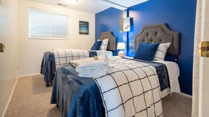 A bedroom with two beds, each with a headboard, a window with blinds, and a wall with a blue accent. - Spacious 3BR Basement Apt | Airport, Work, Ski (North Salt Lake)