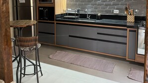 A modern kitchen with a built-in oven, microwave, and stainless steel refrigerator. There are two bar stools and a wooden table. The kitchen has a black countertop and a tiled backsplash. - Sobrado a 700 Metros Praia do Veloso (São Pedro Ilhabela)