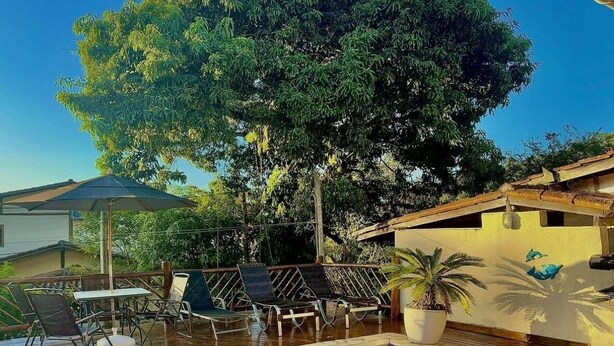 A swimming pool with a tiled edge, surrounded by a wooden deck. There are lounge chairs and a table under a large tree. - Sobrado a 700 Metros Praia do Veloso (São Pedro Ilhabela)