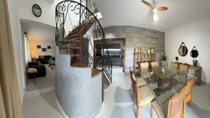 A modern living space with a glass dining table, wooden chairs, a spiral staircase, and a kitchen area. - Sobrado a 700 Metros Praia do Veloso (São Pedro Ilhabela)
