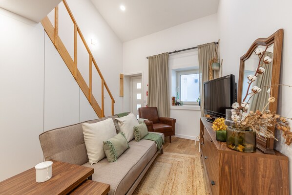 A modern living room with a wooden staircase, a sofa with cushions, a TV, a chair, and a wooden cabinet with decorative items. - Foz Outeiro House 12 (Oporto)